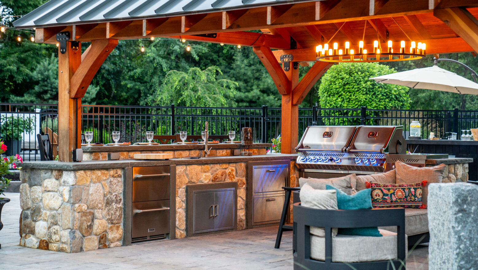 Covered outdoor pavilion kitchen featuring a stone cooking island, dual grill setup, bar seating, and ambient lighting.