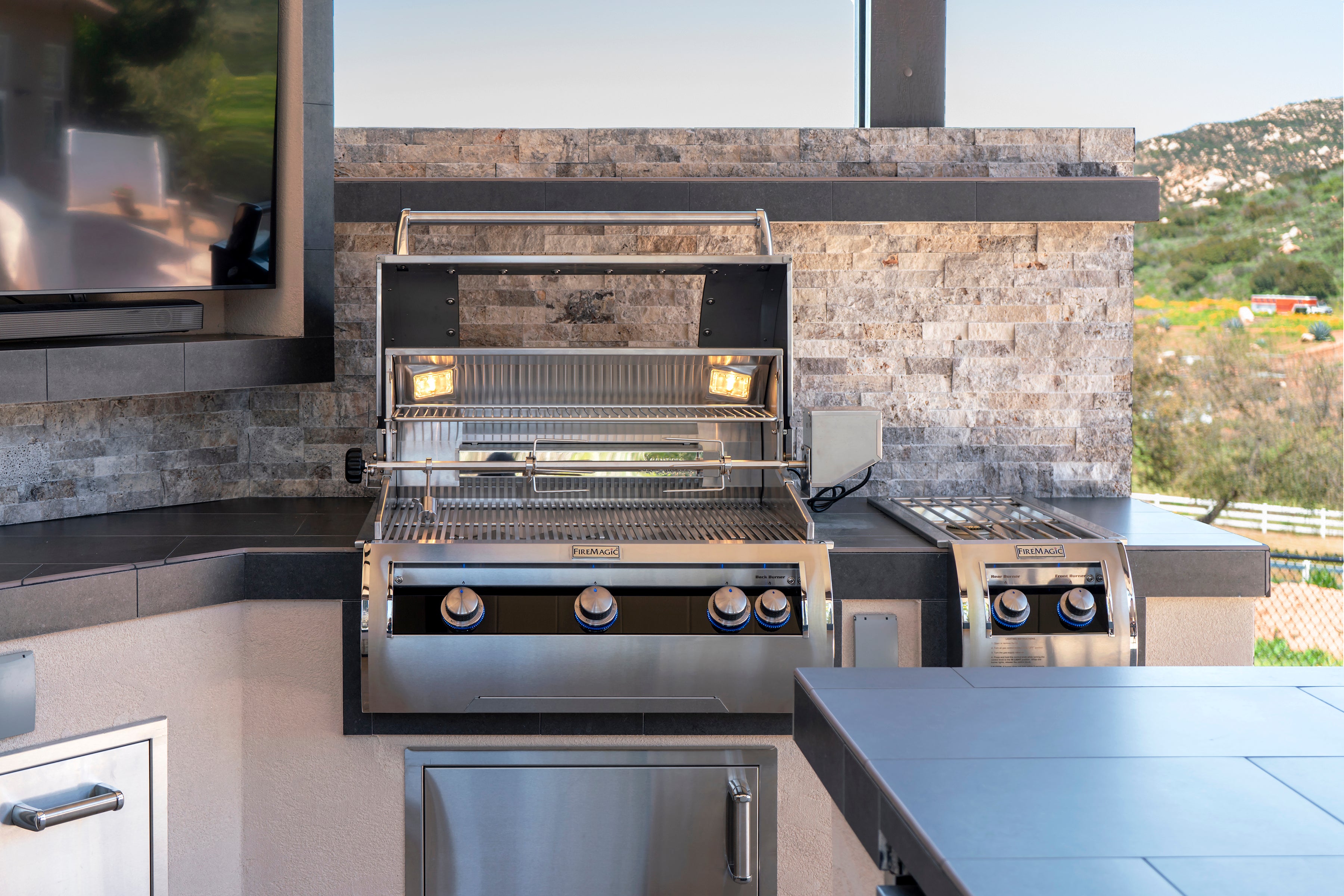 Professional-grade cooking station with rotisserie system, control knobs, and integrated side burner, alongside a built-in TV unit in a stone wall.